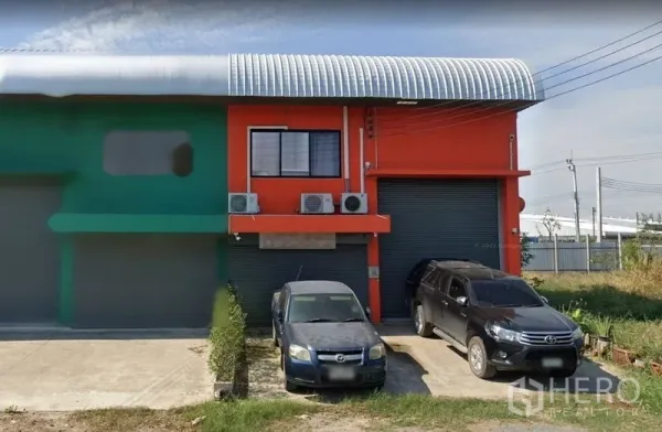 Warehouse for sale in Samut Prakan - Street view of an orange warehouse with twin roller shutter doors, curved metal roof and parking space in front.