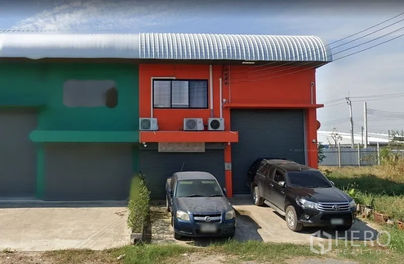 Warehouse for sale in Samut Prakan - Street view of an orange warehouse with twin roller shutter doors, curved metal roof and parking space in front.