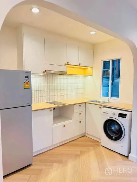 Townhouse for rent in Suan Luang, Bangkok - Modern L-shaped kitchen with white cabinetry, tiled backsplash, refrigerator, sink, and front-load washer.