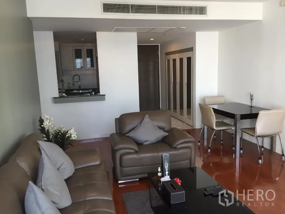 Condominium for rent or sale in Khlong Toei, Bangkok - Open-plan living room with brown leather sofas, dining set and a pass-through kitchen on glossy wood floors.