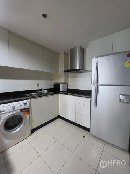 Condominium for rent in Huai Khwang, Bangkok - Modern L-shaped kitchen with white cabinets, black countertops, washer, fridge, and hood.