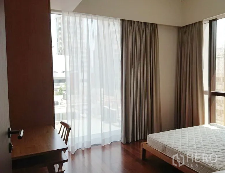 Condominium for sale in Pathum Wan, Bangkok - Corner bedroom with large windows, sheer curtains, a work desk and bed frame.