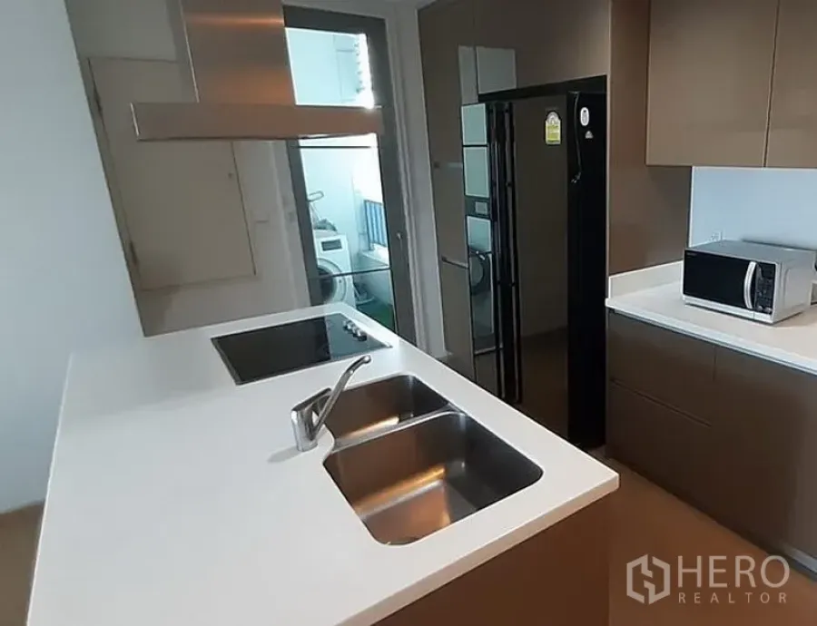 Condominium for rent in Khlong Toei, Bangkok - Modern condo kitchen with white island, double sink, induction hob, and a laundry nook behind a glass door in Khlong Toei.