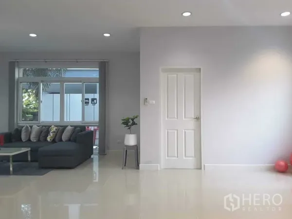 Detached House for rent in Lam Luk Ka, Pathum Thani - Spacious living room with a gray L-shaped sofa beside a large window in neutral tones.
