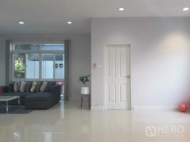 Detached House for rent in Lam Luk Ka, Pathum Thani - Spacious living room with a gray L-shaped sofa beside a large window in neutral tones.