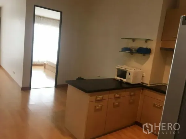 Condominium for rent in Khlong Toei, Bangkok - Compact L-shaped kitchen with black countertops, cabinets, microwave and shelves, facing a bright bedroom.