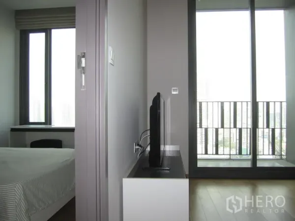 Condominium for rent in Khlong Toei, Bangkok - Sliding glass doors open to a balcony with city view next to a TV stand and adjacent bedroom window.