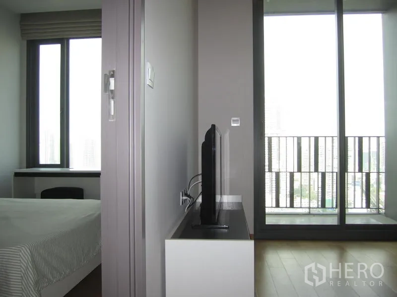 Condominium for rent in Khlong Toei, Bangkok - Sliding glass doors open to a balcony with city view next to a TV stand and adjacent bedroom window.