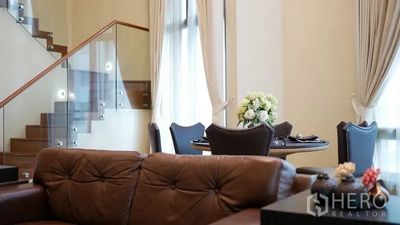 Condominium for rent in Khlong Toei, Bangkok - Living space with a glass-railed staircase, tufted sofa and dining nook with a floral centerpiece.