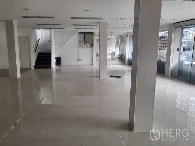 Shophouse for rent in Khlong Toei, Bangkok - Open-plan ground floor of a shophouse with glossy tile flooring, structural columns, a staircase, and wide glass frontage.
