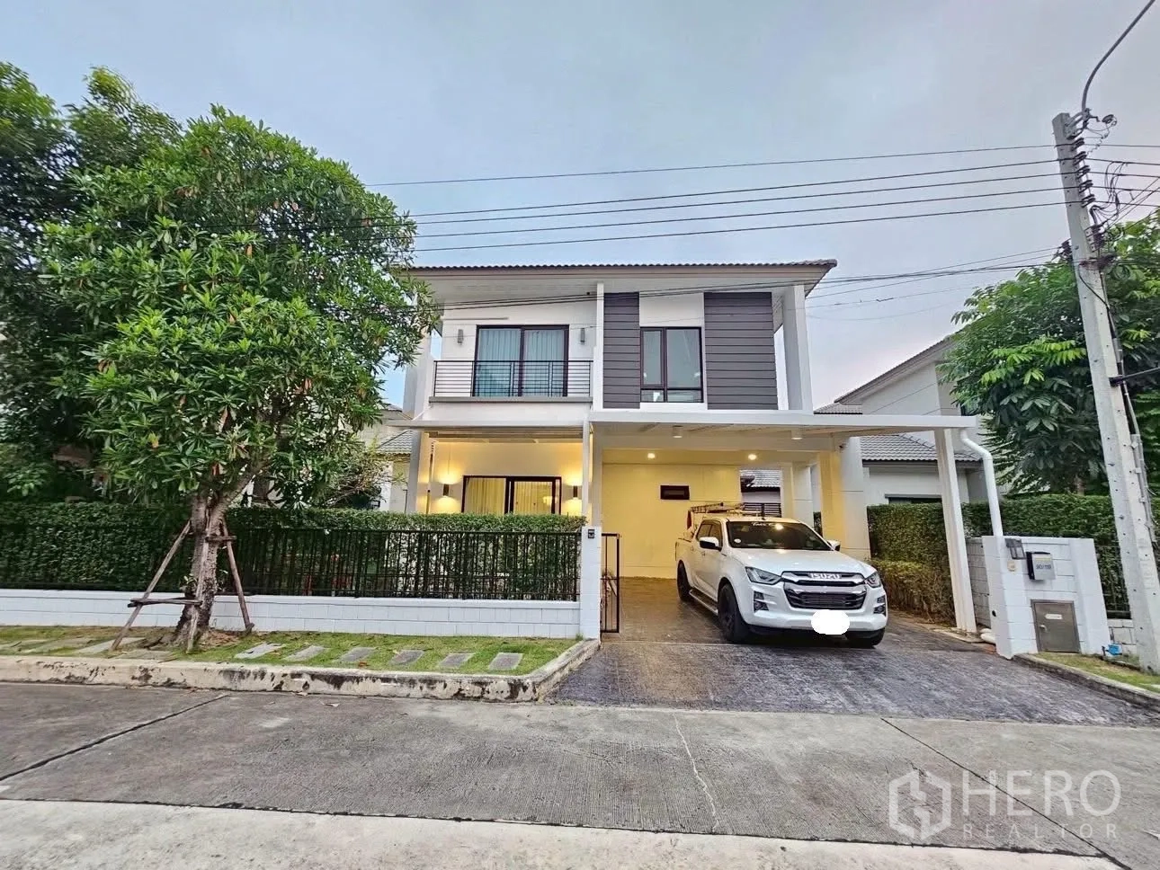 独立式住宅 出售在Bang Phli, Samut Prakan - 两层独栋别墅外观，车棚与铺装车道，草坪和绿篱，社区街景。