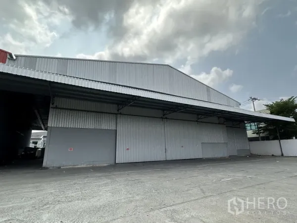 Warehouse for rent in Mueang Samut Prakan, Samut Prakan - Angled exterior of the warehouse with metal awning and open maneuvering space for trucks.