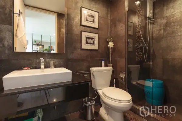 Condominium for rent in Khlong Toei, Bangkok - Modern bathroom with dark tiles, glass-enclosed shower, and floating vanity.