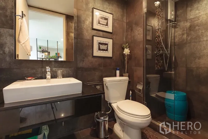 Condominium for rent in Khlong Toei, Bangkok - Modern bathroom with dark tiles, glass-enclosed shower, and floating vanity.