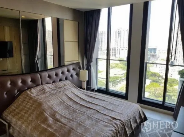 Condominium for rent in Pathum Wan, Bangkok - Corner bedroom with panoramic city views through tall windows and padded headboard bed.