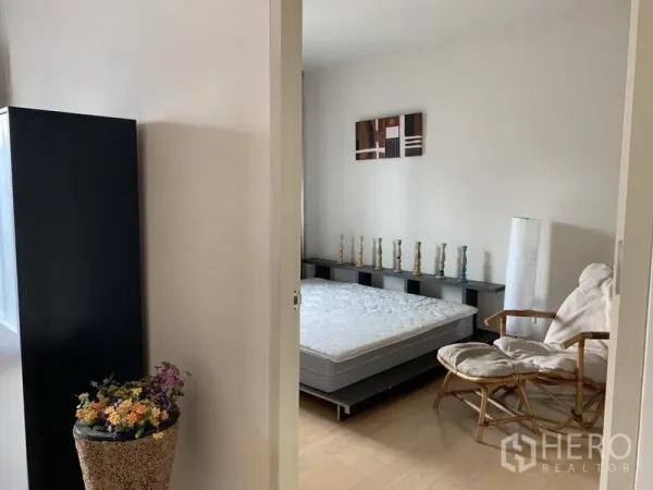 Condominium for rent in Phaya Thai, Bangkok - View into the bedroom showing a mattress, rattan lounge chair, decorative shelf, and neutral décor.