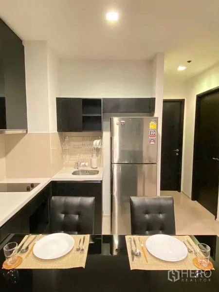 Condominium for rent in Khlong Toei, Bangkok - Compact kitchen with stainless refrigerator, sink, cabinetry and a dining table set for two.