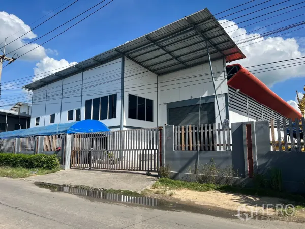 Warehouse for sale in Lam Luk Ka, Pathum Thani - Street view of a modern white warehouse with metal roof, roller shutter, and secure gated fence in Lam Luk Ka.