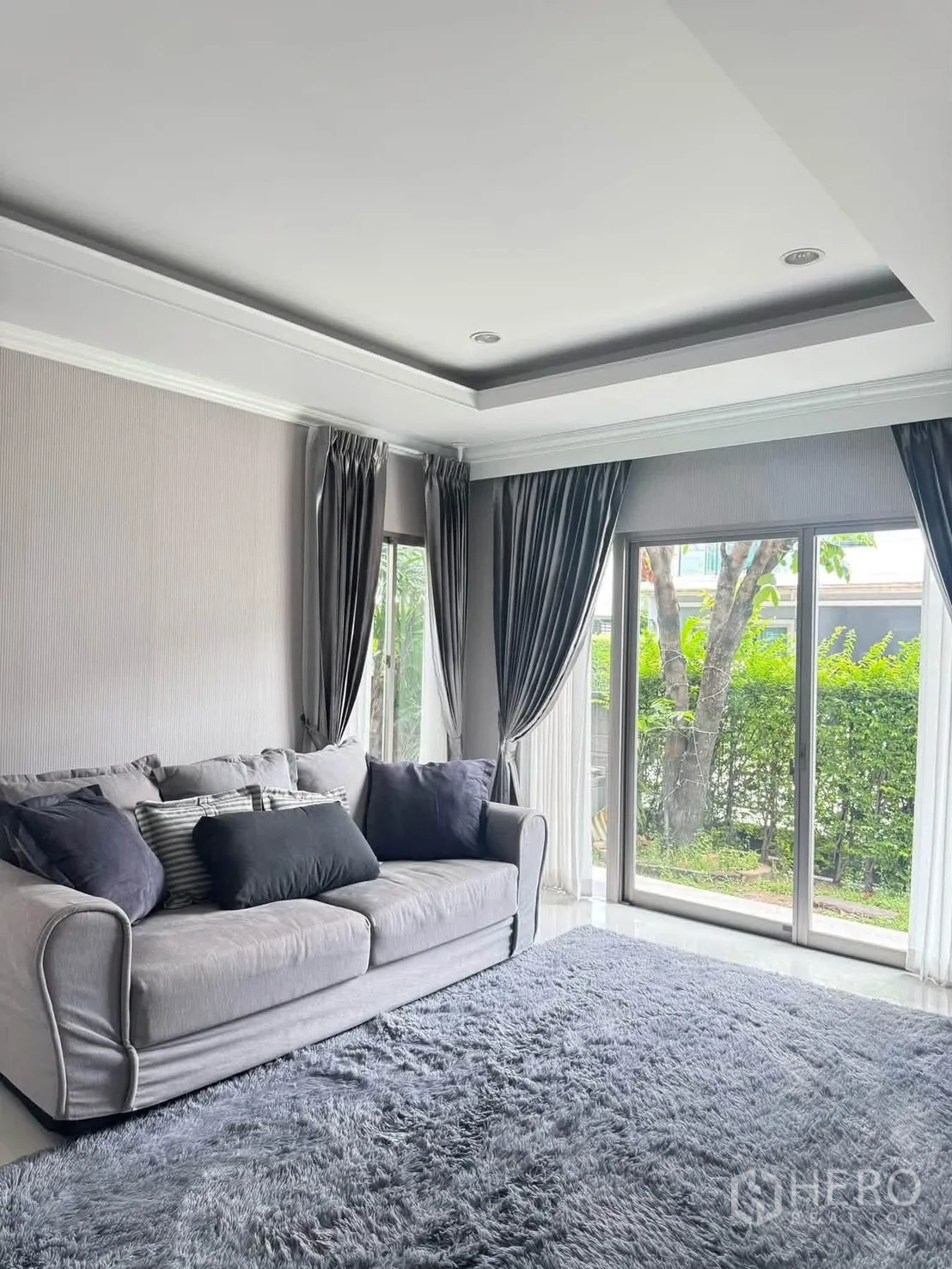 半独立式住宅 for rent or sale in Suan Luang, Bangkok - 客厅放置灰色沙发与厚地毯，落地玻璃面向绿色景观。