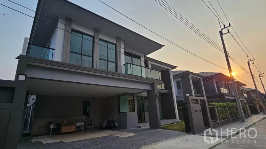 Detached House for rent or sale in Prawet, Bangkok - Modern two-storey detached house facade with glass balcony and covered carport on a quiet street at sunset.