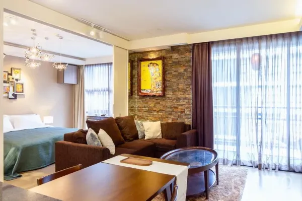 Condominium for rent in Watthana, Bangkok - Bright living area with sectional sofa, stone feature wall, sheer curtains, and bedroom separated by sliding partition.