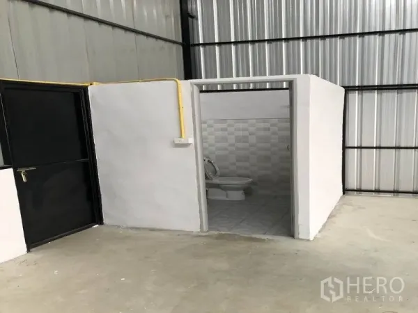 Warehouse for rent in Pathum Thani - In-warehouse restroom with tiled walls beside a black metal door and yellow utility conduit.