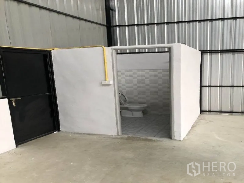 Warehouse for rent in Pathum Thani - In-warehouse restroom with tiled walls beside a black metal door and yellow utility conduit.