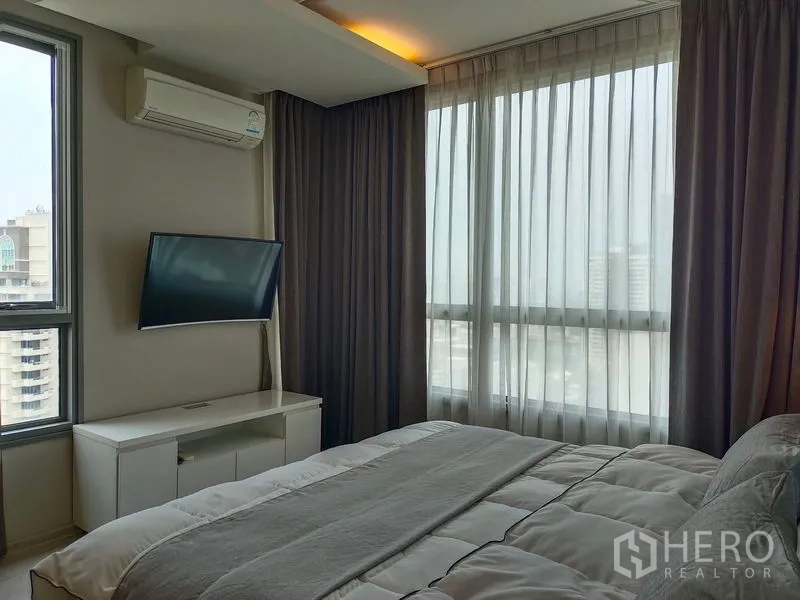 Condominium for rent in Watthana, Bangkok - Corner bedroom with wall-mounted TV, sheer curtains and panoramic windows.