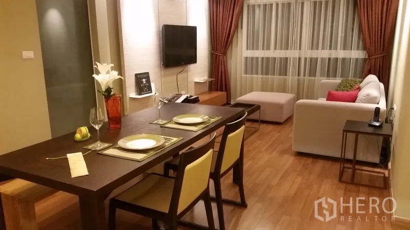 Condominium for rent in Khlong San, Bangkok - Open-plan living and dining room with wood floors, sofa, TV wall and a large curtained window.