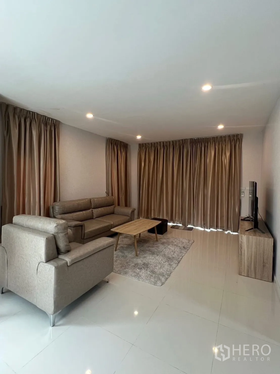 บ้าน ให้เช่าในBang Phli, Samut Prakan - ห้องนั่งเล่นตอนกลางวัน ผ้าม่านเต็มความสูง ชุดโซฟาสีเทา ทีวี และพื้นกระเบื้องเงา