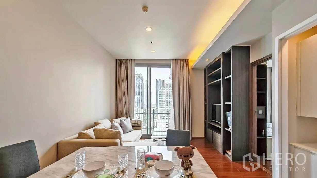 公寓 出租在Watthana, Bangkok - 开放式客餐厅，米色转角沙发、内置电视柜、木地板，落地推拉门通向观景阳台俯瞰曼谷天际线。