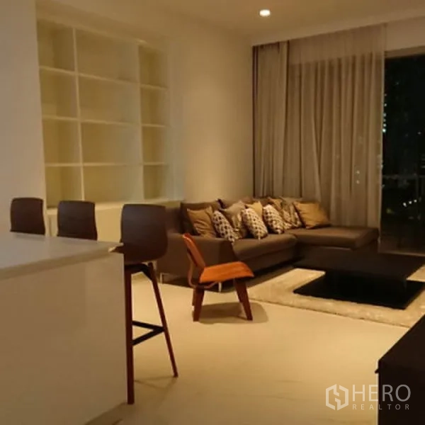 公寓 出租在Pathum Wan, Bangkok - 温馨客厅配L形沙发、嵌入式收纳与落地窗帘，连接阳台。