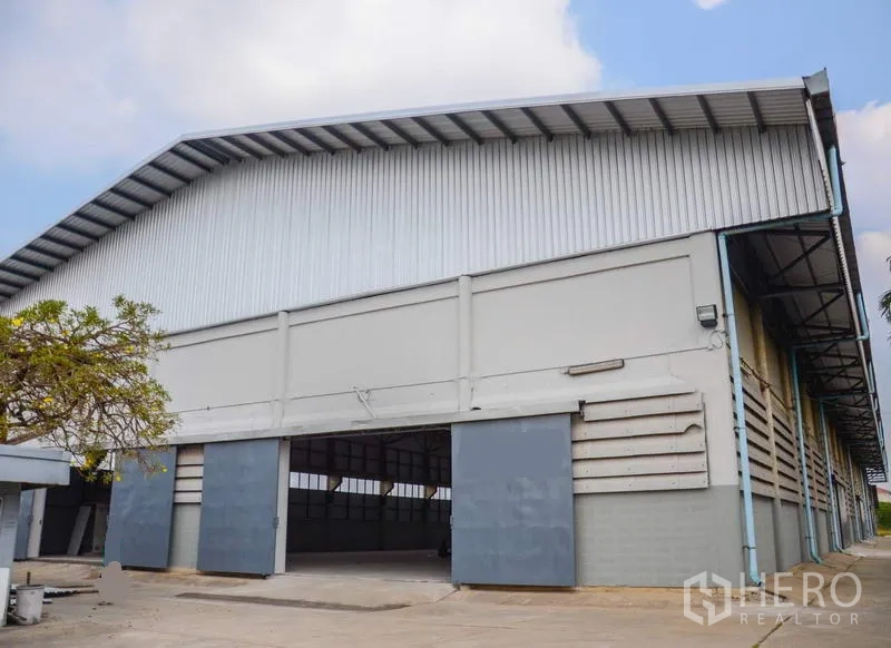 工厂 出租在Pathum Thani - 高挑屋顶的仓库外立面，超宽推拉门与混凝土装卸区。