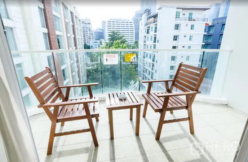 Condominium for rent in Khlong Toei, Bangkok - Balcony with two wooden chairs and a small table overlooking low-rise buildings and greenery.