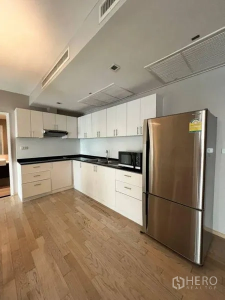 Condominium for rent in Khlong Toei, Bangkok - Modern L-shaped kitchen with white cabinets, black countertop and stainless refrigerator.