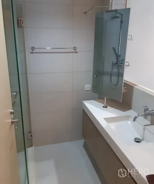 Condominium for rent in Khlong Toei, Bangkok - Contemporary bathroom with glass-enclosed shower, long vanity counter, and neutral wall tiles.