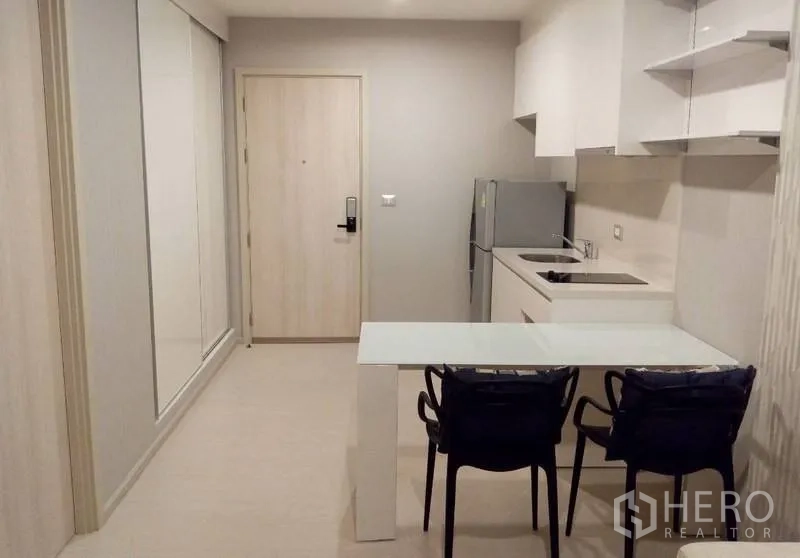 Condominium for rent in Khlong Toei, Bangkok - Compact kitchenette with white cabinets, refrigerator, cooktop and a glass dining table near the entry door.