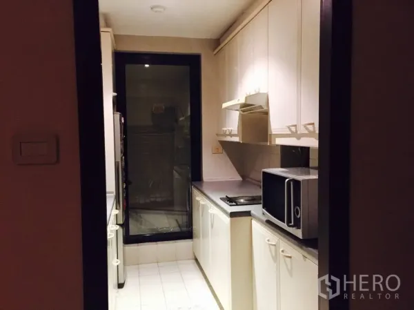 Condominium for rent in Sathon, Bangkok - Compact galley kitchen with white cabinets, cooktop, range hood, microwave and a glass door.