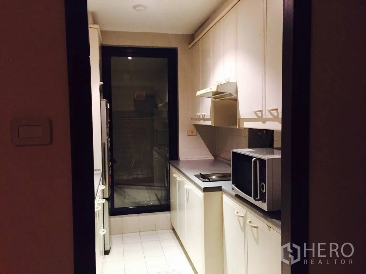 Condominium for rent in Sathon, Bangkok - Compact galley kitchen with white cabinets, cooktop, range hood, microwave and a glass door.