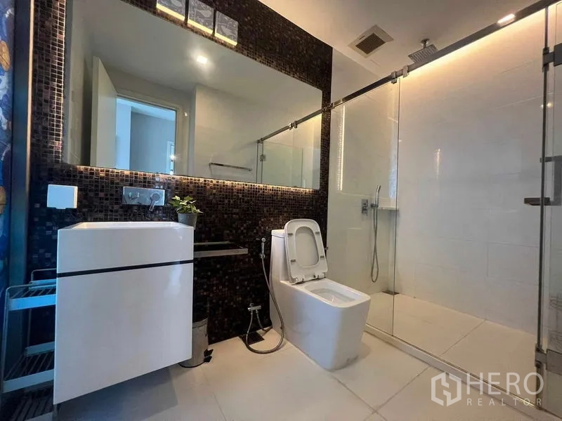 Condominium for rent in Khlong Toei, Bangkok - Modern bathroom featuring a large mirror, glass-enclosed shower and mosaic accent wall.