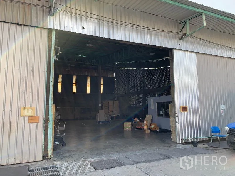 Warehouse for rent in Mueang Pathum Thani, Pathum Thani - Wide metal sliding doors open to a high‑ceiling warehouse with concrete flooring, skylight windows and a small internal office.