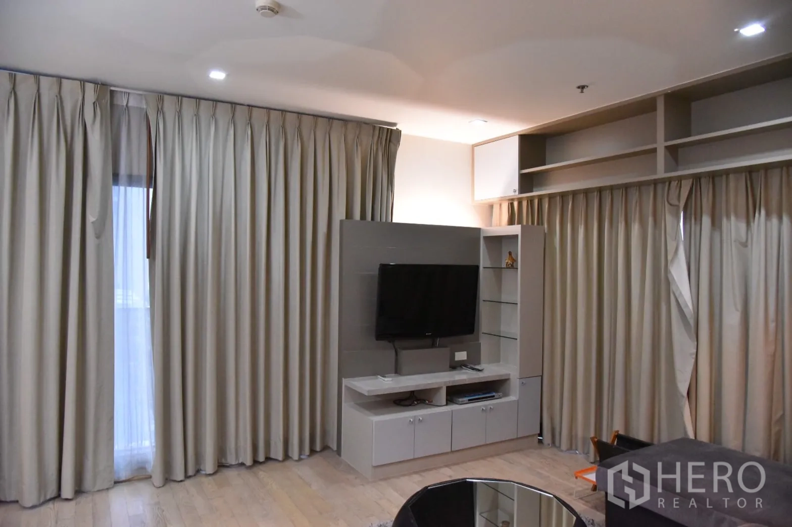 Condominium for rent or sale in Khlong Toei, Bangkok - Living room with built-in TV wall, shelving, light wood flooring and full-height curtains.