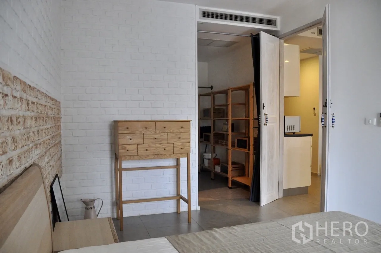 Condominium for rent in Bangkok - Bedroom corner with white brick walls, tall wooden chest and walk-in shelving next to the kitchen entrance.