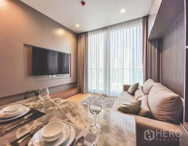 公寓 出租在Bang Rak, Bangkok - 客厅配沙发与电视墙，整面高窗帘，旁为大理石餐桌。