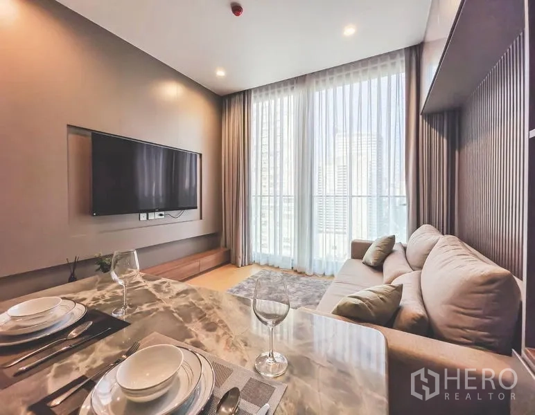 公寓 出租在Bang Rak, Bangkok - 客厅配沙发与电视墙，整面高窗帘，旁为大理石餐桌。