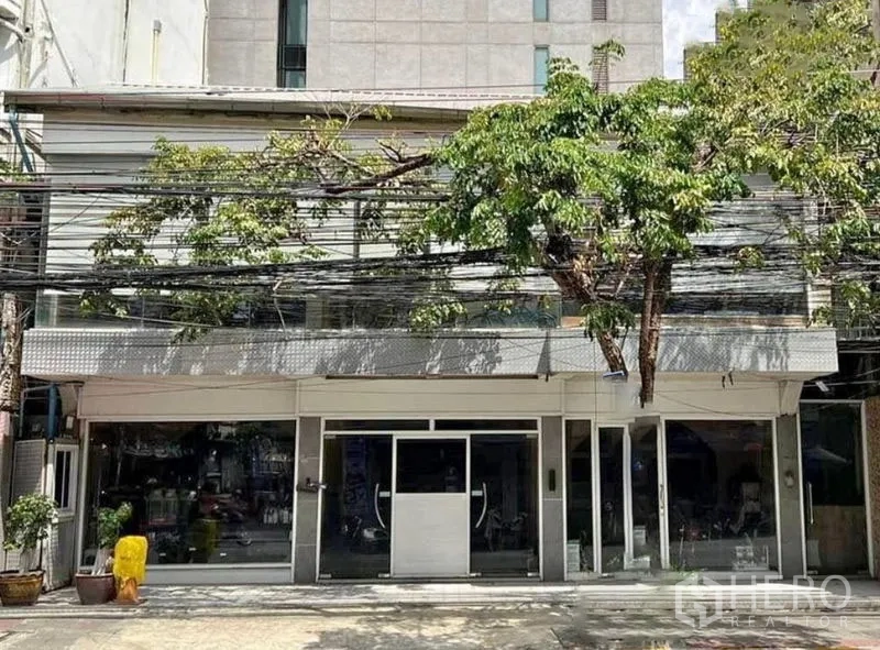 Shophouse for rent in Watthana, Bangkok - Street-facing two-storey shophouse with wide glass storefront shaded by mature trees.