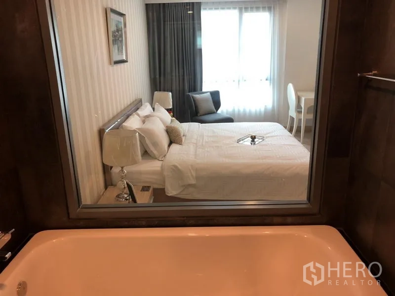 公寓 出租在Watthana, Bangkok - 从浴室望向主卧，设浸泡浴缸、玻璃隔断、床与大窗的明亮空间。