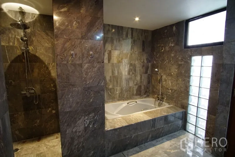 Apartment for rent in Watthana, Bangkok - Luxury bathroom clad in stone tiles with separate rain shower and a sunken bathtub beside a glass-block window.