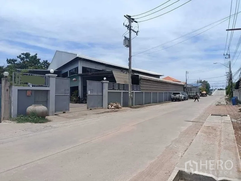 Warehouse for sale in Samut Sakhon - Street view of a gated warehouse compound with tall perimeter wall, power lines and a wide concrete road in Mueang Samut Sakhon.