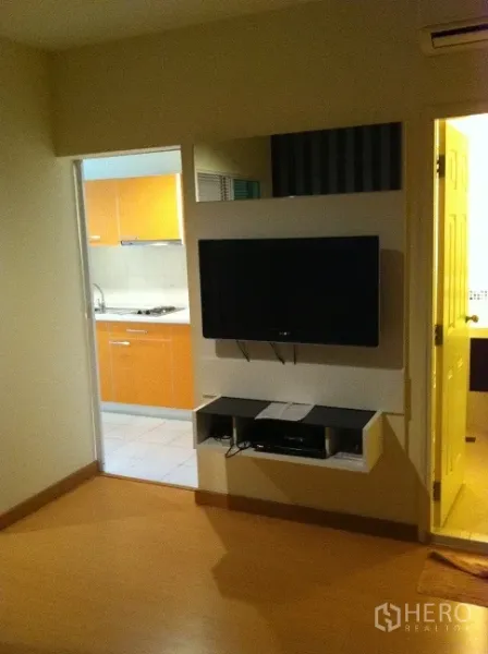 公寓 出租在Watthana, Bangkok - 墙挂电视与置物架，对面为客厅；旁侧通向带橙色柜体的独立厨房